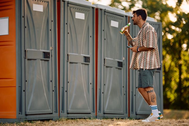 Person drinking beverage while standing near row of portable toilets at outdoor event
