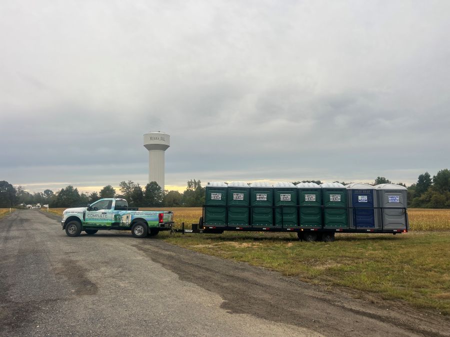 Portable toilet trailer hauled by service truck near field and water tower in Deptford, NJ
